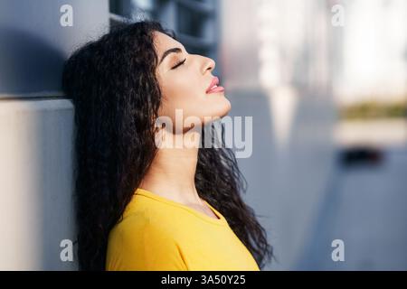 Arab woman with curly hair wearing a yellow shirt stands against a wall on a city street, eyes closed in a thoughtful moment. The urban outdoor setting and copy space support lifestyle, fashion, and travel storytelling. A contemporary portrait with a calm mood. Stock Photo