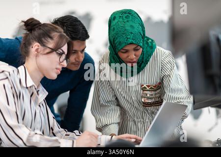 Multiethnic startup team in a modern open plan office brainstorming around a laptop. Arabian woman wearing a hijab participates in the discussion with colleagues in a collaborative work setting. Suitable for business, innovation, and diverse workplace campaigns. Stock Photo