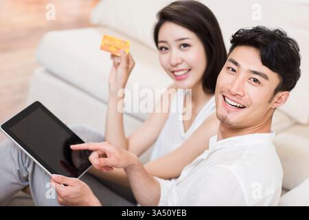 Cheerful Chinese couple shopping online on a digital tablet at home. They share a relaxed moment, illustrating e commerce and togetherness in a contemporary living space. Stock Photo