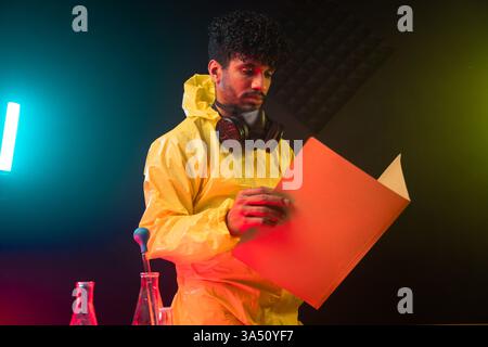 Serious Hispanic male chemist in a hazmat suit and gas mask reads a document in a dark laboratory beside pipettes and flasks. Ideal for science, research, lab safety and industrial communications. Stock Photo