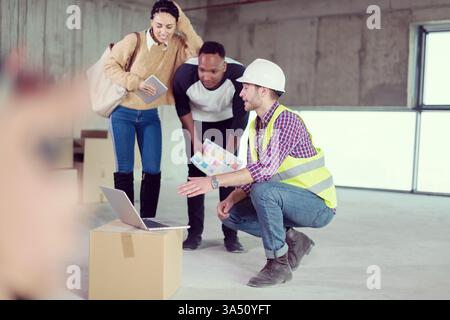 Architect engineer uses a laptop to show house design plans to a young multiethnic couple on a construction site. A client-facing moment in a modern building project. Useful for architecture, home design, and new home development topics. Stock Photo