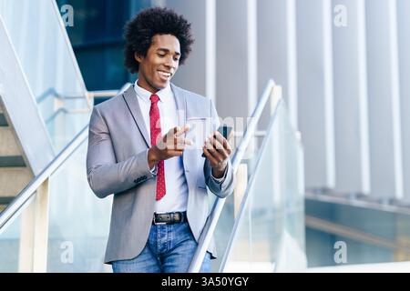 Black businessman uses his smartphone beside an office building. Confident professional portrait highlighting mobile technology in the workplace. Ideal for business, technology, and corporate lifestyle imagery. Stock Photo