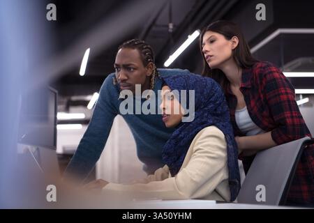 A diverse team of software developers collaborates in a modern startup office. They work together at a laptop, writing code and building software in a professional, energetic setting. Stock Photo