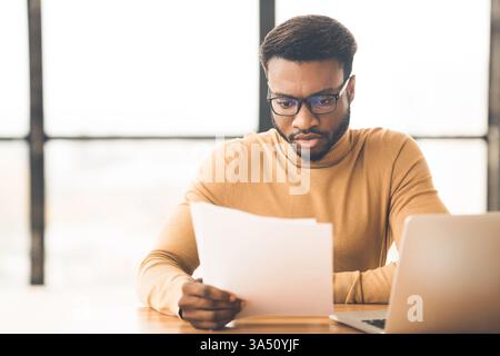 Concentrated Black businessman studying financial reports at a modern office with copy space. A professional, corporate scene ideal for finance, business, and workplace communications. Stock Photo