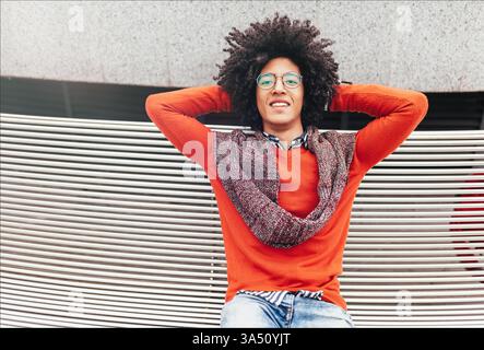Handsome young Egyptian man with curly hair sits on a bench outdoors in a bright orange sweater and jeans, smiling at the camera. This confident, student-style portrait works well for features on study abroad, campus life, and multicultural experiences. Stock Photo