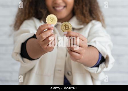 Smiling African American woman extends her arms to display cryptocurrency coins against a wall. This vibrant fintech image works well for stories about crypto, blockchain, and digital currencies. Ideal for marketing fintech, investment concepts, and modern finance materials. Stock Photo