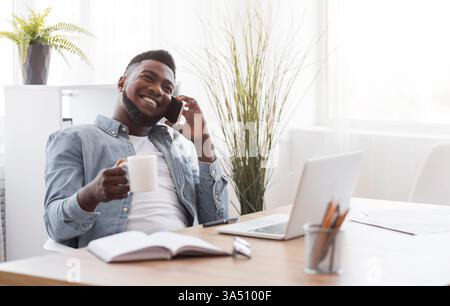Cheerful African American professional on a coffee break, talking on the phone and holding a hot drink in the office. This workplace lifestyle image conveys positive energy, collaboration, and modern business culture. Stock Photo