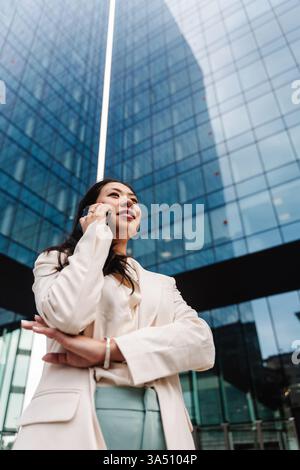 Professional Asian woman on a phone call in a modern corporate office conveys focus and confidence. The sleek blazer and smartphone create a strong business portrait suitable for corporate, finance, and leadership visuals. Ideal for messages about ambition, success, and urban work life in business communications. Stock Photo