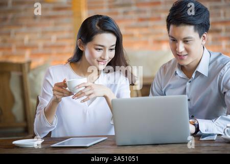 Smiling Chinese man uses a laptop while a Chinese woman holds a cup of coffee at a cafe table. This collaborative scene suits business, teamwork, and cafe culture imagery. Ideal for digital work, online collaboration, and modern office vibes. Stock Photo