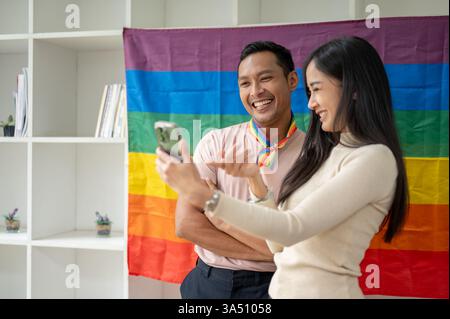 Cheerful Asian woman taking a selfie with a gay friend standing against an LGBT flag at home. A joyful moment of friendship, inclusion, and Pride. Perfect for LGBTQ+ lifestyle, celebration, and social media visuals. Stock Photo