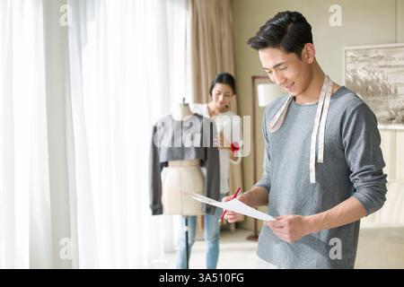 Two Chinese fashion designers collaborating in a clothing design studio. The image shows sketching, tape measures and a mannequin, capturing creativity and small business fashion work. Great for articles about fashion design, collaboration, and entrepreneurship. Stock Photo