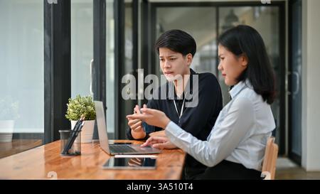 Confused Asian male and female office workers pointing at laptop screen having problem with their project while sitting together at table in cafe Stock Photo