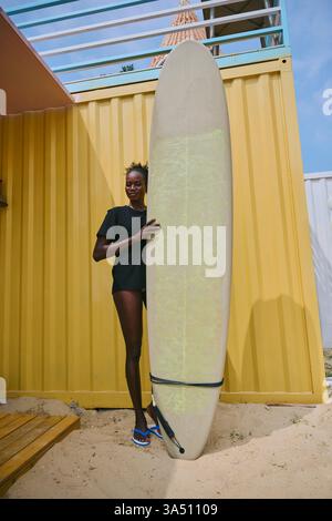 Smiling Black woman standing with a surfboard at a sunny beach bar. This coastal lifestyle image suits travel, sports and summer campaigns. Perfect for beachwear, surf culture, and active lifestyle branding. Stock Photo