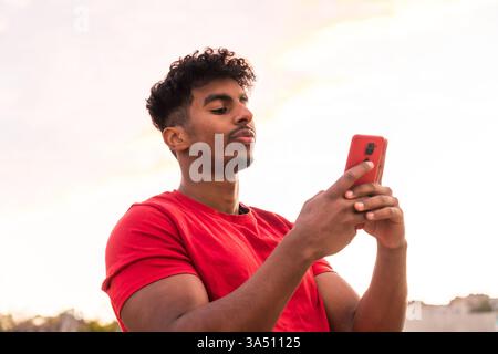 Arab young man in a red t-shirt types a message at sunset in a city street. Casual and confident, he uses a smartphone in an urban outdoor setting, perfect for technology and lifestyle campaigns. Stock Photo