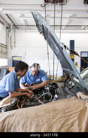 Black female and Caucasian male mechanics inspect a car engine in a repair shop. They wear uniforms in an industrial setting, conveying teamwork and skilled auto repair. A suitable choice for automotive maintenance and shop culture visuals. Stock Photo