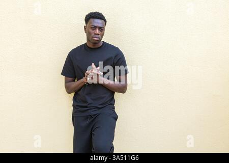 Concentrated young Black man in casual wear standing against a pale yellow wall on a street, looking at the camera with a focused expression. A contemporary street style portrait suitable for lifestyle, fashion or urban campaigns. Stock Photo
