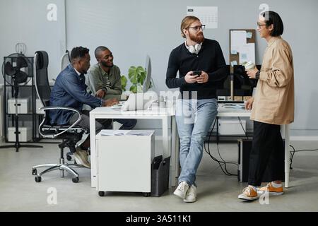 A diverse group of young program managers chat during a break in a modern office, while two colleagues work at computers nearby. This contemporary workplace scene suits business, teamwork, and corporate culture themes. Stock Photo