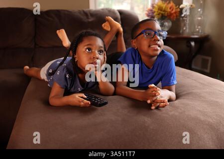 Black girl leans her face on her hand while using a remote control to watch TV, with her brother lying down on the couch nearby. A warm home life moment that highlights sibling bonding and everyday family entertainment. Stock Photo