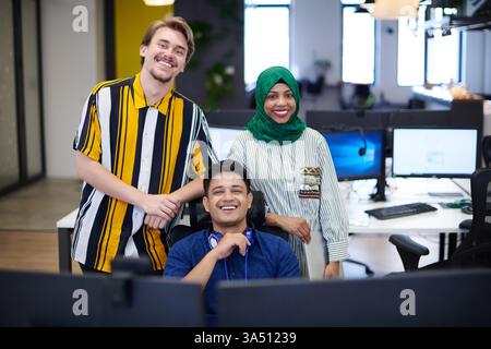 Diverse startup team in a modern open-plan office brainstorming session. An Arabian woman wearing a hijab collaborates with colleagues around a laptop and desktop computer, highlighting teamwork and technology. Stock Photo