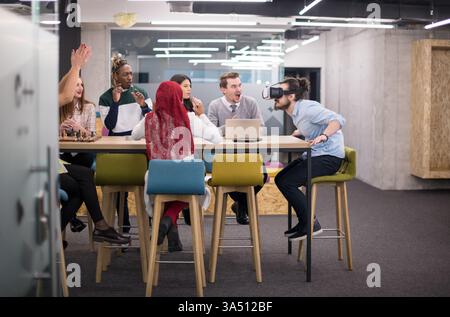 Cheerful mixed race male developer wearing a VR headset, experiencing virtual reality as colleagues cheer during a break. The office tech scene highlights teamwork, innovation, and immersive technology. Stock Photo
