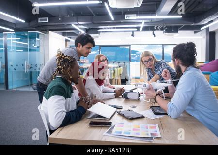 Diverse group of business people sit around a table with laptops and documents while discussing drone technology and VR glasses for business advancement. The scene conveys teamwork, innovation, and strategic planning in a modern office. Ideal for technology, startup, and future-focused business visuals. Stock Photo