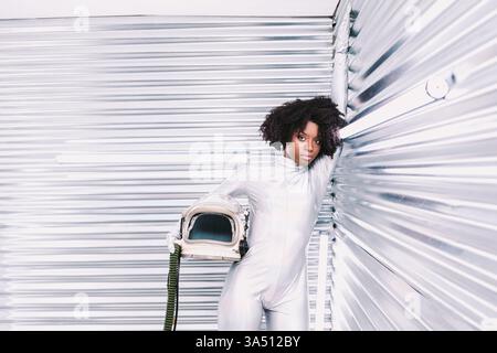 Serious Hispanic female astronaut with Afro wearing a spacesuit holds a helmet and leans on a silver wall. The light room adds a futuristic, professional vibe for space and science imagery. Useful for STEM, women in science, and space career projects with copy space. Stock Photo