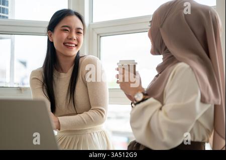 Cheerful Asian woman chats with a Muslim friend in a cafe. They hold coffee at a table by the window with a laptop, suggesting a casual meet and remote work. Stock Photo