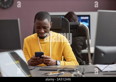 Portrait of a Black software developer at an office desk using a smartphone. The high tech workspace highlights coding, data, and cybersecurity themes. Great for tech, IT, software development, and digital innovation campaigns with copy space. Stock Photo