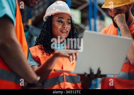 Black female worker wit curly hair wearing hard hat and safety vest discussing inventory on laptop with colleagues standing in warehouse during work Stock Photo