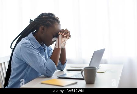 Depressed Black businesswoman seated at a desk near a window, touching her forehead in apparent headache or stress. This image conveys workplace pressure, challenge, and fatigue in a professional setting. Suitable for business, entrepreneurship, and mental health themes. Stock Photo