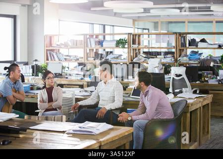 Diverse group of office workers from different backgrounds chat and share ideas in a modern office. They sit around a table, highlighting teamwork and creative collaboration. This image suits workplace culture, collaboration, and design-focused business content. Stock Photo