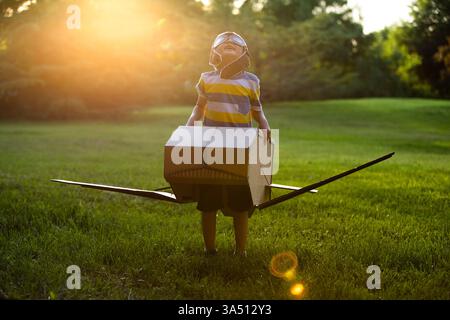 Smiling Chinese boy wearing an airplane cardboard box costume stands in a park at sunset. A playful, imaginative scene ideal for family, childhood, and summer activity campaigns with outdoor warmth. Great for lifestyle and travel content emphasizing creativity and fun. Stock Photo