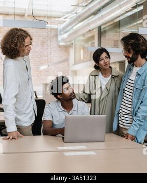Diverse group of colleagues collaborating in a modern office, engaged in discussion around a table with a laptop. The scene emphasizes teamwork, professional dialogue, and a collaborative work culture. Great for business, diversity, and office communication themes. Stock Photo