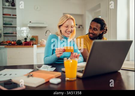 Cheerful multicultural couple at home using a laptop and credit card for online shopping. This modern ecommerce scene is perfect for content about online shopping, digital payments, and family finance. Stock Photo