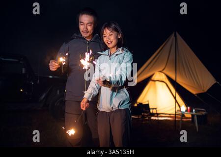 Happy Chinese couple outdoors camping, holding sparklers under a night sky. They are surrounded by nature, sharing a moment of romance and togetherness. Great for travel, holidays and lifestyle visuals or romantic couple imagery. Stock Photo