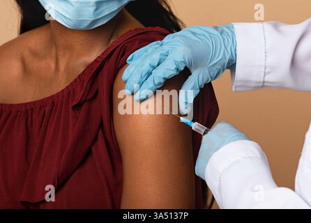Unrecognizable doctor in workwear and gloves administers a vaccine to a lady in a beige studio background. Ideal for healthcare, vaccination campaigns and medical promotion visuals. Stock Photo