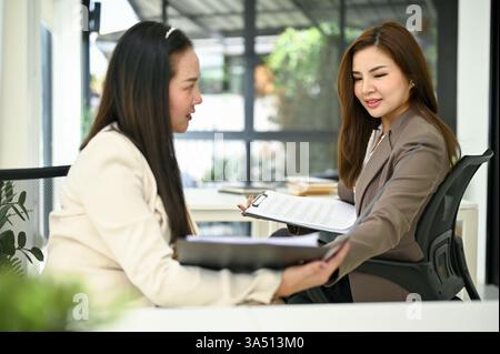 Professional millennial Asian businesswoman collaborating with a colleague in a modern office, sharing ideas during a meeting. This scene is ideal for topics on women in business, teamwork and workplace collaboration. It highlights focus, professionalism and productive discussion in a corporate environment. Stock Photo