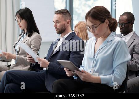 Group of intercultural brokers in formalwear reviewing documents at a conference. Diverse professionals collaborate in a modern boardroom with papers and tablets. Stock Photo