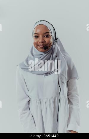 Portrait of an African Muslim businesswoman wearing a hijab and headset, appearing as a customer service representative in a modern office. She smiles while assisting a customer online, projecting professionalism and support in a call center setting. Stock Photo