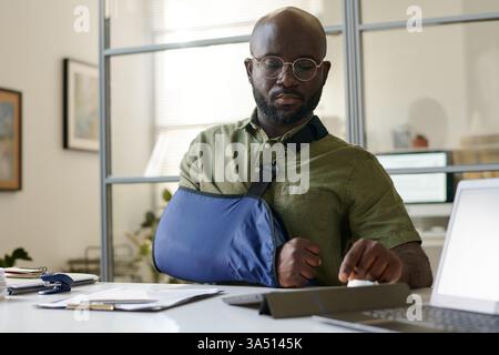 An African American man sits in a social service office, using a tablet to manage an injury claim. The scene conveys professional assistance and digital workflows in an indoor public service environment. Suitable for topics like client service, insurance, and administration. Stock Photo