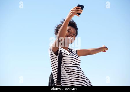 Portrait of a beautiful young African American woman taking a selfie outdoors against a clear sky. She points away from the camera with a playful gesture, adding a dynamic element to lifestyle and social media visuals. Perfect for outdoor content, travel, and personal branding. Stock Photo