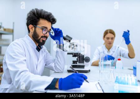Smiling Indian male scientist making notes of his investigation while sitting at table near caucasian female colleague working on liquid chemicals ... Stock Photo