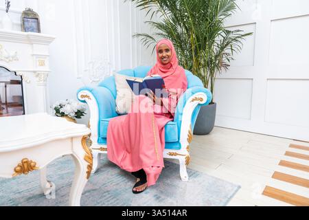 Smiling Muslim woman in pink abaya and hijab sits at home holding a Quran. The intimate reading moment is set on a sofa in a comfortable living room, emphasizing Islamic spirituality and daily life. Suitable for faith-based content, home lifestyle, and relaxation themes. Stock Photo