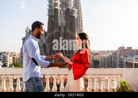 Multiracial couple dating on a rooftop balcony in Barcelona near the Sagrada Familia. They share a romantic moment over drinks, capturing happiness and togetherness against an iconic city backdrop. Perfect for travel, romance, and destination marketing. Stock Photo
