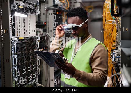 Waist-up portrait of a technician in a data center, repairing servers and inspecting computer parts. The image highlights hardware maintenance, server rooms, and IT support work. Ideal for articles about data centers, network infrastructure, and technical services. Stock Photo
