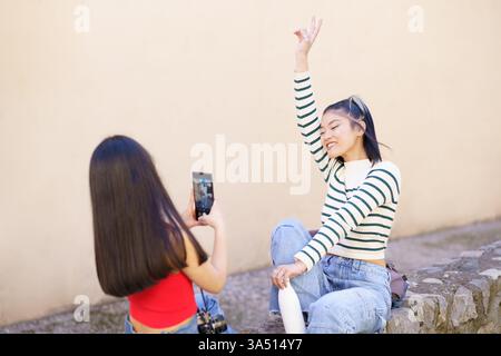 Two joyful Asian women tourists sitting on a stone border and taking photos with a smartphone in a town street. Ideal for travel, sightseeing, and vacation imagery featuring friends exploring urban destinations. Great for content about travel, photography, and social sharing. Stock Photo