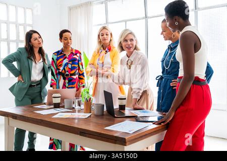 Diverse group of smiling businesswomen collaborating around a table with a laptop and documents. They discuss plans and brainstorm in a modern office setting. Stock Photo