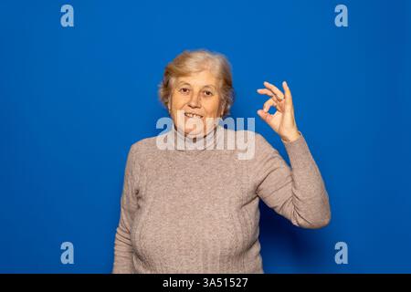 Close-up photo of smiling Caucasian senior retired woman standing casually, showing Ok gesture symbol, looking at camera isolated over blue color Stock Photo