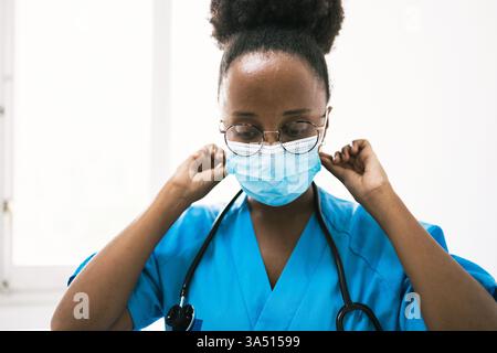Front-on portrait of a Black female physician in a blue medical uniform, adjusting a protective mask with stethoscope at the ready. This professional healthcare image works for medical, hospital and clinical contexts, conveying competence and safety. Perfect for campaigns about patient care, medical services and diversity in medicine. Stock Photo