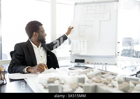 Successful mixed-race man architect working on an estate construction project in the office. The professional setting highlights design, architecture, and construction planning with blueprints and plans. Ideal for architecture firms, engineering, real estate marketing, and construction campaigns. Stock Photo